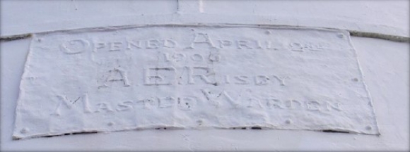 Photograph showing a white plaque fitted to outer side of lighthouse tower. Plaque reads "Opened April 2nd 1906 A.E.R ISBY Master Warden" 