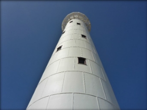 Photograph showing tall, white lighthouse tower consisting of cast iron panels bolted together. Several windows are fitted into the tower at varying heights. 