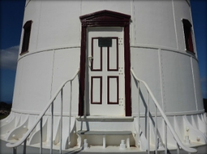 Photograph showing tower door fitted to base of white lighthouse tower. Stairs and railing lead to door. 