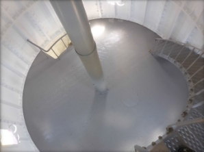 Photograph showing round, grey floor fitted within lighthouse tower. A grey weight tube is fitted to centre of floor, and a grey spiral staircase leading to a floor above.  