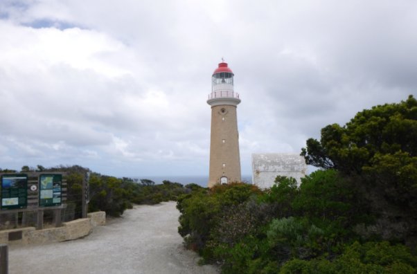 Photograph of a dirt path leading past green vegetation and a white, arched shed to a stone lighthouse tower with red dome.