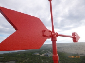 Photograph showing red weathervane sitting atop lighthouse dome. Various ex-keepers cottages can be seen in distance below.