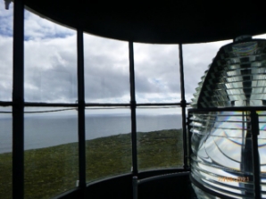 Photograph showing internal of lantern house with prismatic lens sitting behind curved glazing panes. Grey sea can be seen through panes. 