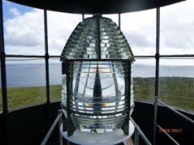 Photograph showing prismatic lighthouse lens sitting behind curved glazing panes. Sea and sky can be seen through panes.