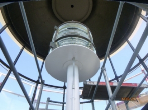 Photograph showing lighthouse lens sitting atop a steel pedestal inside a curved lantern room.