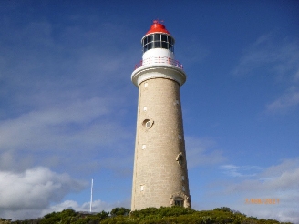 Photograph showing stone lighthouse tower with red dome and white lantern house against blue sky.