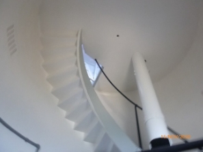 Photograph showing underside of curved staircase leading up to circular floor above. A weight tube is fitted to the underside of the floor above. 