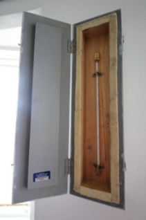 Photograph showing thermometer sitting in timber case, fitted to wall. 