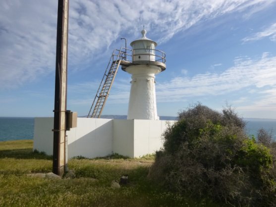 Photograph showing small white lighthouse tower with external balcony ladder surrounded by solid white fence against a blue sky. 