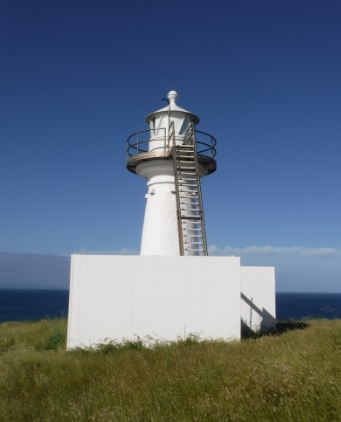 Photograph showing small white lighthouse tower with external balcony ladder surrounded by solid white fence against a blue sky. 