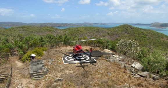 Photograph showing a red helicopter perched on a helipad, surrounded by green vegetation. 