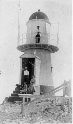 Black and white photograph of small lighthouse tower. A person stands on the balcony above, and three persons and a dog stands beside the front door of the tower.  
