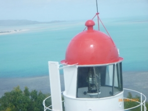 Photograph showing circular lantern house with red dome roof. A prismatic lens can be seen sitting inside the lanternroom. A blue ocean can be seen behind.