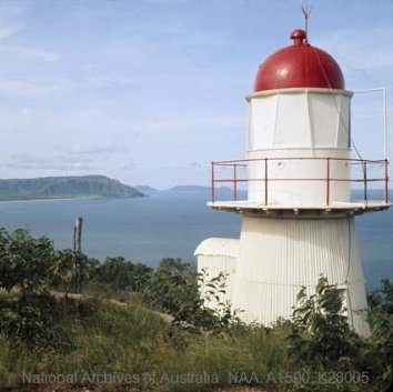 Photograph showing small white lighthouse with red dome roof amongst green vegetation, overlooking the sea.