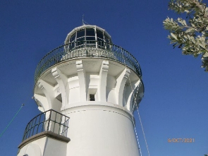 Photograph showing white lighthouse tower with external staircase and large lantern room and balcony supported by stone brackets. 