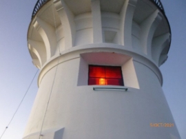 Photograph showing lighthouse tower with rectangular window set into the walls. The window has red glass panes which are illuminated with a light. 